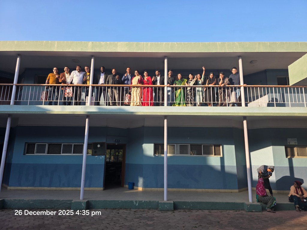 Us, ex-students, standing on a balcony, smiling and waving, with a school building in the background.